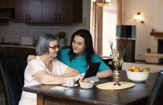 Hoitaja opastaa ikäihmistä tabletin käyttämiseen keittiön pöydän ääressä.