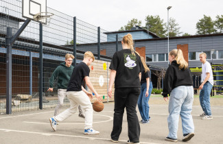 Ryhmä oppilaita pelaa koripalloa koulun pihalla.