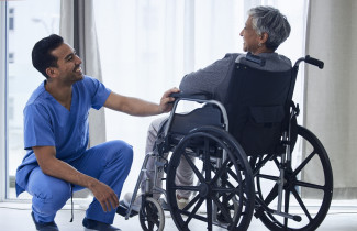 Siniasuinen hoitaja kyykyssä pyörätuoliasiakkaan edessä