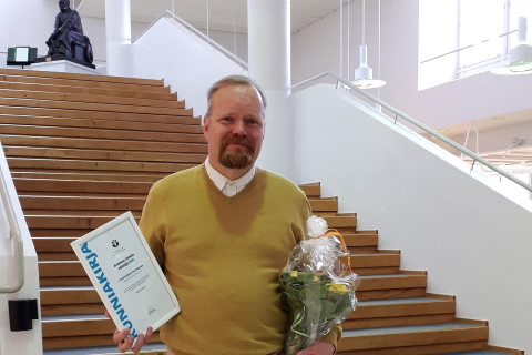 Mies seisoo portaiden edessä toisessa kädessään kunniakirja ja toisessa kukkakimppu.