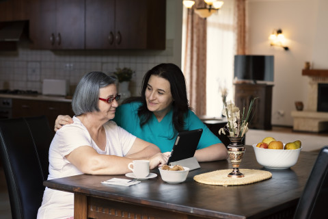 Hoitaja opastaa ikäihmistä tabletin käyttämiseen keittiön pöydän ääressä.