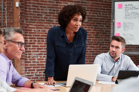 Nainen johtaa puhetta, ihmisiä pöydän ympärillä.