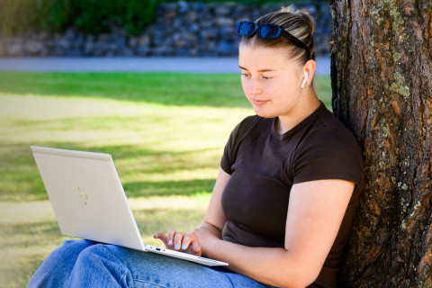 opiskelija iskumassa puun juurella ja tekemässä töitä kannettavalla tietokoneella