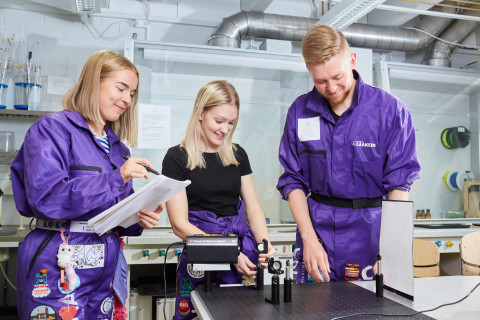 Kolme fotoniikan opiskelijaa valolaboratoriossa violetit haalarit yllään