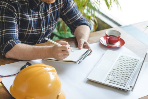 Henkilö kirjoittaa kannettavan tietokoneen äärellä lehtiöön vierellään pöydälle laskettu turvakypärä.