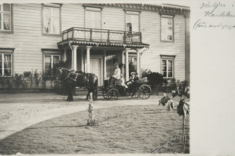 Kaksi henkilöä hevosen vetämissä rattaissa kartanorakennuksen edessä.