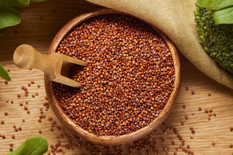 Top view of red quinoa in a wooden bowl with a spatula next to a beige napkin and green vegetables.