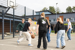 Ryhmä oppilaita pelaa koripalloa koulun pihalla.