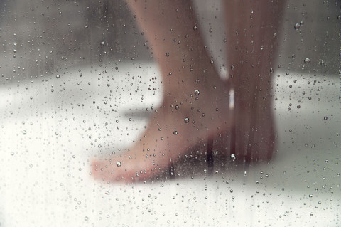Female Bare feet are positioned on a white shower floor, with a glass door displaying water droplets, creating a soft focus on the background.