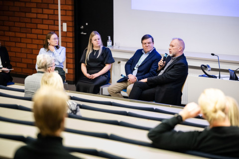 Neljä panelistia istuu luentosalin etuosassa kasvot ylesiöön päin.