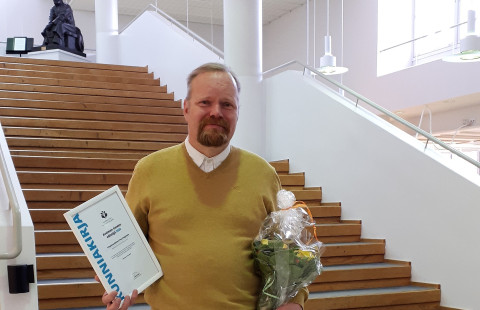 Mies seisoo portaiden edessä toisessa kädessään kunniakirja ja toisessa kukkakimppu.