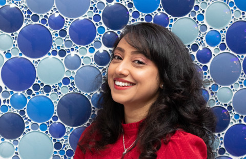 Nainen punaisessa paidassa hymyilee kameralle takanaan sinisistä lasiympyröistä muodostuva tausta.