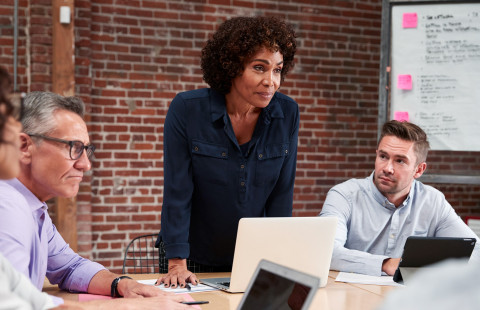 Nainen johtaa puhetta, ihmisiä pöydän ympärillä.