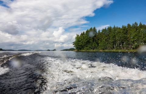 Veneen perävana järven pinnassa. Taustalla saari ja pilvinen taivas.