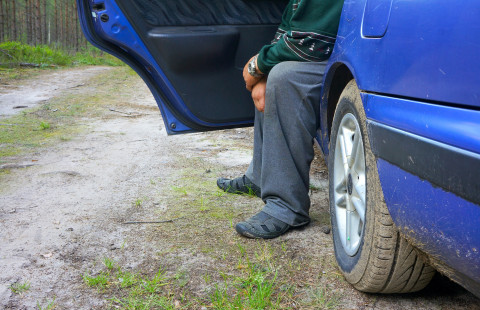 Henkilö istuu auton etupenkillä jalat auton ulkopuolella.