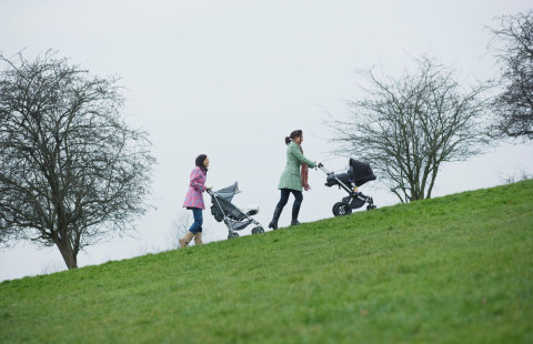 Kaksi äitiä työntää peräkkäin lastenvaunuja ruohikkoiseen rinteeseen taustalla lehdettömiä puita.