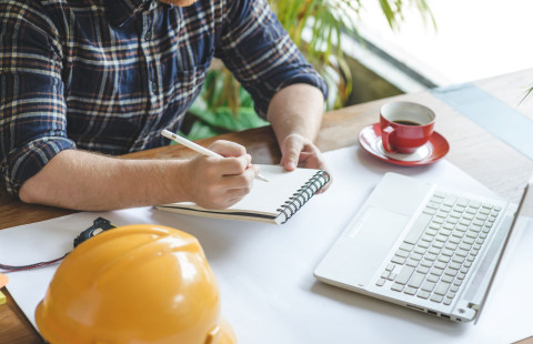 Henkilö kirjoittaa kannettavan tietokoneen äärellä lehtiöön vierellään pöydälle laskettu turvakypärä.