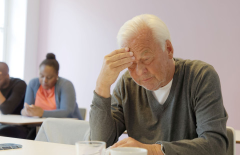 Ikääntynyt henkilö nojaa otsaa käteensä luokkahuoneen päydän ääressä.