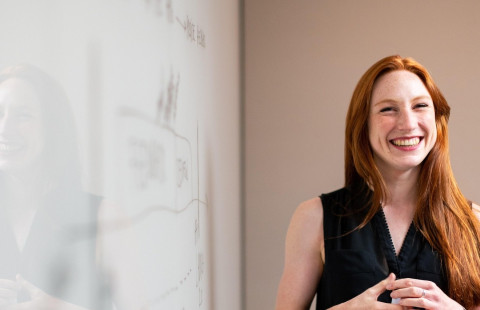 Punahiuksinen hymyilevä nainen seisoo valkotaulun edessä