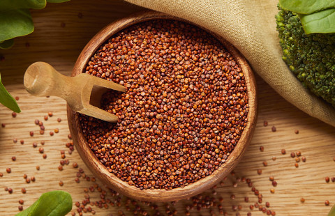 Top view of red quinoa in a wooden bowl with a spatula next to a beige napkin and green vegetables.