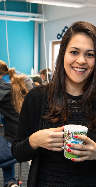 Young woman looking at the camera smiling and holding a coffee cup, young men working with their laptops in the background.